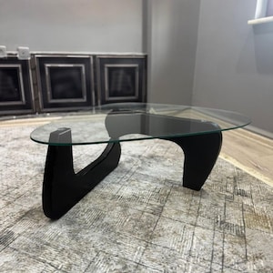 Puede incluir: Una mesa de centro con un tablero de cristal transparente de forma orgánica, apoyado sobre una base de madera maciza negra. La base consta de dos patas curvas interconectadas que proporcionan una base estable para la superficie de cristal.