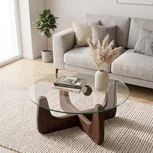 May include: A round coffee table with a clear glass top and a dark brown wooden base featuring four curved legs that intersect in the center. The table holds two stacked books titled Nordic Interiors and Form and Function, a small vase, and a gray object.