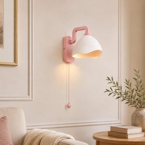 May include: A wall-mounted lamp featuring a pink rectangular base and a curved arm supporting a white, bell-shaped shade with a textured surface. A pink pull cord with a small matching pendant hangs from the base, emitting a warm light.