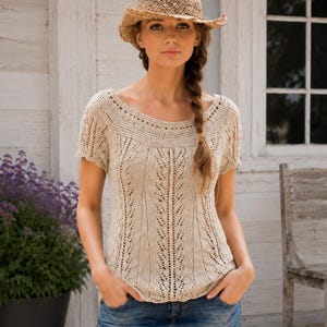 May include: A beige short-sleeved knit top featuring an intricate openwork lace pattern and a wide rounded neckline, paired with a blue denim mini skirt and a woven straw hat with a wide brim and a dark brown band around the crown.