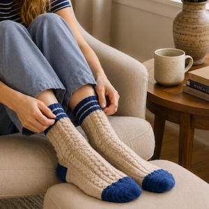 Puede incluir: Un par de calcetines de punto trenzado en color beige con detalles en azul oscuro en las puntas, talones y puños. Los puños presentan finas rayas horizontales blancas. Estos calcetines de media caña tienen un patrón de punto texturizado.