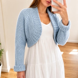 Puede incluir: Un bolero de punto azul claro con mangas largas, puños doblados y un cuello ancho de canalé. La prenda se lleva sobre un vestido blanco sencillo sin mangas. La textura del tejido es visible en toda la tela de color azul claro.