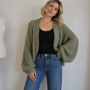 May include: A sage green open-front cardigan with a chunky knit texture, long balloon sleeves, and ribbed trim along the collar and cuffs. The cardigan is worn over a simple black tank top and paired with classic blue denim jeans.