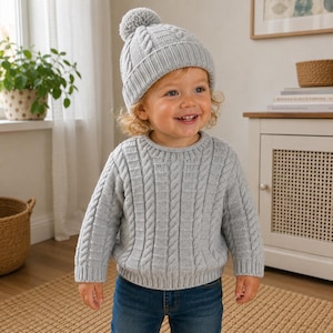 May include: A light grey long-sleeve cable knit jumper and a matching knit beanie with a pom-pom on top. The jumper features a vertical cable pattern and a ribbed crew neckline, while the beanie has a ribbed cuff and a textured knit design.