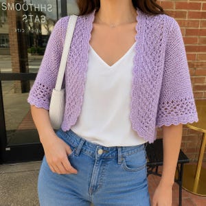May include: A light purple open-front crochet cardigan with short sleeves and a scalloped edge, worn over a plain white camisole and blue denim jeans. A white shoulder bag with a thin strap is positioned at the side of the torso.