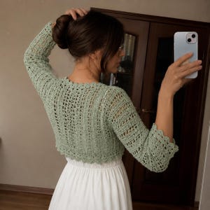 May include: A sage green crochet bolero featuring an intricate openwork pattern, long sleeves, and a scalloped edge along the neckline, cuffs, and hem. The garment is worn over a plain white dress, showcasing the detailed texture of the yarn work.