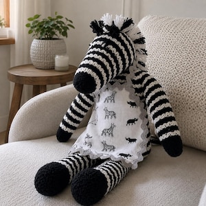 Puede incluir: Un peluche de cebra tejido a ganchillo con rayas blancas y negras, una melena de hilo blanco y un delantal de tela blanca con un estampado negro repetitivo de cebras y rinocerontes con un borde festoneado gris alrededor.