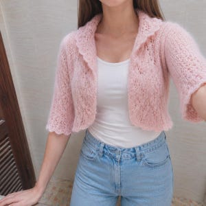 May include: A light pink, open-front bolero cardigan featuring a delicate, airy knit pattern with a scalloped collar and elbow-length sleeves. The garment is worn over a simple white tank top and light blue denim jeans, showcasing a soft texture.