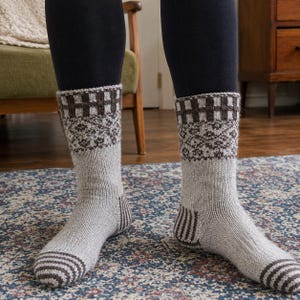 Puede incluir: Un par de calcetines de punto a media pantorrilla con una base gris claro jaspeado y detalles en marrón oscuro. El diseño incluye un patrón de rayas marrón oscuro en la puntera y el talón, una banda geométrica detallada y un patrón de rejilla.