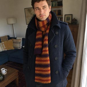 Könnte beinhalten: Ein dunkelblauer zweireihiger Mantel mit hohem Kragen, getragen mit einem langen, gestrickten Schal mit horizontalen Streifen in den Farben Dunkelviolett, gebranntes Orange und Goldgelb. Der Schal hat eine texturierte, wollartige Optik.