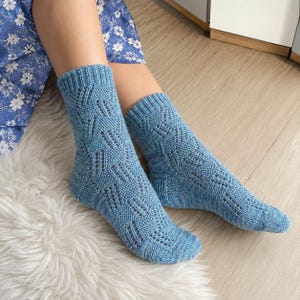 May include: A pair of mid-calf length socks in a light blue hue, featuring a detailed knit pattern with vertical eyelet columns and a ribbed cuff. The socks are crafted with a textured stitch design that runs from the toe up to the ankle.