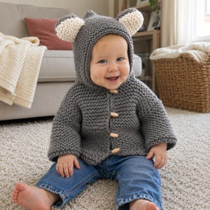 May include: A dark gray knitted hooded cardigan featuring two cream-colored bear ears on top of the hood and four light-colored wooden toggle buttons down the front. The garment is worn with blue denim jeans, creating a textured and cozy appearance.