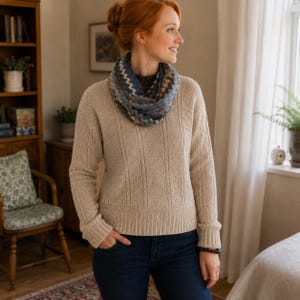 May include: A beige cable-knit sweater with a ribbed hem and cuffs, paired with a blue and white patterned infinity scarf. The sweater features vertical textured panels and a diamond stitch design, worn over dark blue denim jeans.