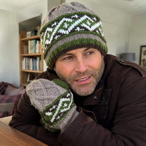 Puede incluir: Un conjunto de gorro y manoplas de punto con una base de color marrón claro y una banda decorativa con patrones geométricos en marrón oscuro, verde bosque y blanco. El gorro tiene un borde acanalado y las manoplas muestran un diseño de trenzas.
