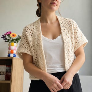 Op de afbeelding: Een crèmekleurig, gehaakt vest met korte mouwen, voorzien van een open voorzijde en een gestructureerd, herhalend vierkant patroon. Het kledingstuk is gemaakt van lichtgewicht garen en toont een verfijnde, luchtige steek over het geheel.