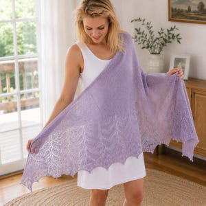 Puede incluir: Un chal de punto de color morado claro con un delicado patrón de encaje con motivos de hojas a lo largo del borde inferior festoneado. El chal está colocado sobre un sencillo vestido blanco sin mangas, mostrando su textura y color.