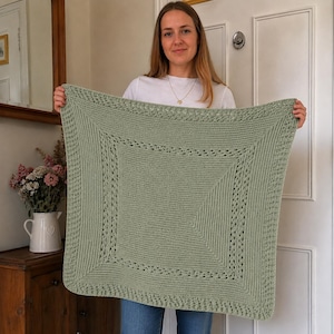 Könnte beinhalten: Eine quadratische, salbeigrüne Strickdecke mit einem zentralen glatt rechts gestrickten Feld, das von einem strukturierten Rand mit einem sich wiederholenden Lochmuster umgeben ist. Die Decke hat eine einheitliche hellgrüne Farbe und ein geometrisches Design.