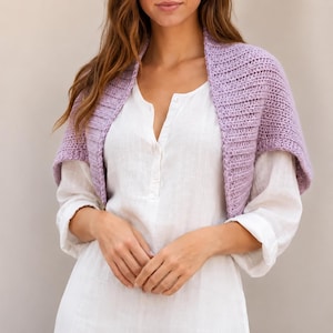 May include: A light purple crochet shrug featuring a textured ribbed pattern, worn over a white long-sleeved shirt with a button-down placket. The shrug has an open front design with short sleeves that drape over the shoulders and upper arms.
