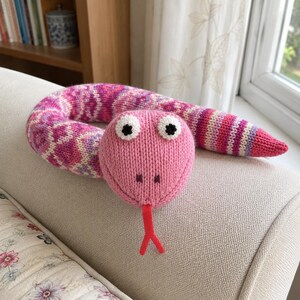 May include: A knitted snake toy featuring a solid pink head with white and black eyes, a red forked tongue, and a long body patterned with pink, purple, and white geometric shapes and stripes. The soft plush toy has a textured yarn construction.