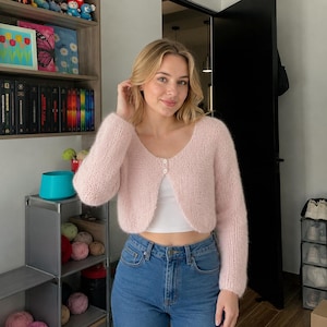 May include: A light pink, long-sleeved, cropped cardigan made of a fuzzy, soft-textured knit fabric. The garment features a rounded neckline and is fastened at the top with two small, round, white buttons. It is worn over a plain white tank top.
