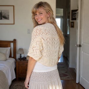 May include: A cream-colored, short-sleeved knit bolero featuring an intricate openwork lace pattern with chevron and floral motifs. The garment is cropped at the waist and worn over a plain white top, paired with a textured, light-colored skirt.