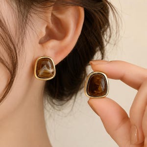 May include: A pair of stud earrings featuring a smooth, rounded, irregular triangular shape. Each earring has a polished gold-tone metal border surrounding a glossy, marbled brown and cream resin centre with a swirling, organic pattern.