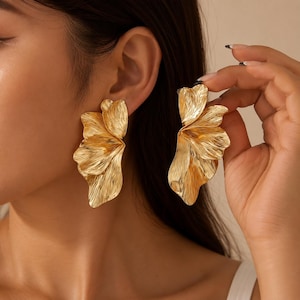 May include: A pair of gold-toned earrings featuring a layered, petal-like design with a textured, metallic finish. The earrings are shaped like abstract, organic floral forms with multiple overlapping segments that create a sense of depth and movement.