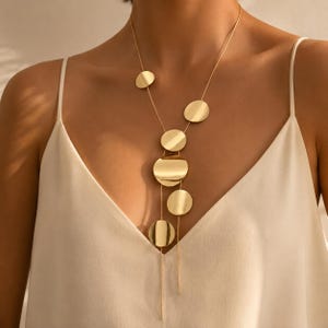 Puede incluir: Un collar de tono dorado que presenta una serie de discos metálicos circulares, lisos y planos, de tamaños variados, suspendidos de cadenas finas y delicadas. Los discos están dispuestos en un patrón vertical asimétrico que cae por el centro del pecho.