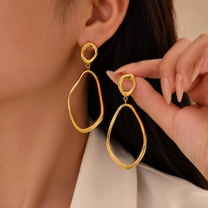 Peut inclure: Une paire de boucles d'oreilles pendantes dorées composées d'un petit clou circulaire irrégulier relié à une forme organique plus grande en goutte d'eau ouverte. Le métal présente une finition lisse et polie avec une surface réfléchissante.