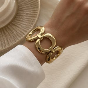 Könnte beinhalten: Ein goldfarbenes Armband bestehend aus einer Reihe großer, polierter, ineinandergreifender ovaler und kreisförmiger Glieder. Das Metall hat eine hochglänzende, reflektierende Oberfläche, die ein markantes, durchgehendes Kettendesign bildet.