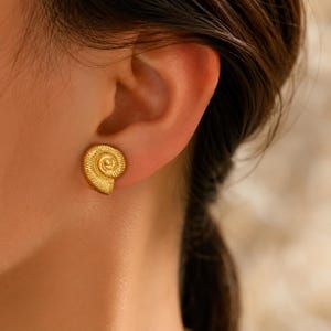 Puede incluir: Un pendiente de botón dorado con forma de concha de nautilo. El pendiente presenta una superficie texturizada en espiral con finas líneas radiantes que imitan los patrones de crecimiento naturales de una concha, sujeto al lóbulo.