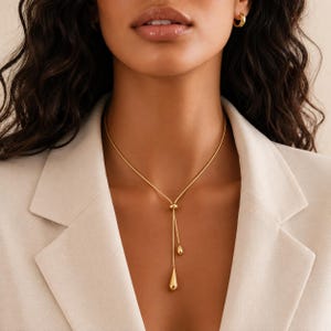 Op de afbeelding: Een goudkleurige lariat-ketting met een dunne ketting die door een kleine ronde verbinding loopt en eindigt in twee hangende druppelvormige hangers van verschillende groottes. De ketting wordt gedragen over een lichtgekleurde blazer.
