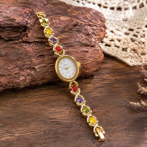 Può includere: Un orologio al quarzo color oro con quadrante bianco e lancette nere. Il cinturino presenta un design a maglie intrecciate incastonate con pietre preziose ovali multicolori, tra cui pietre rosse, viola, gialle e verdi disposte in modo simmetrico.