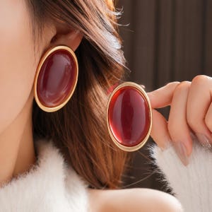 Peut inclure: Une paire de boucles d'oreilles ovales dotées d'une pierre cabochon rouge foncé lisse sertie dans un cadre en métal doré poli. Les boucles d'oreilles présentent un design arrondi et substantiel qui souligne le contraste entre le centre rouge et le bord métallique.