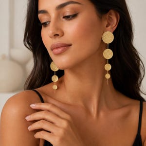 Peut inclure: Une paire de boucles d'oreilles pendantes dorées composées d'un alignement vertical de trois disques circulaires de tailles différentes. Chaque disque présente une surface texturée et gravée. Les boucles sont suspendues par une fine chaîne.
