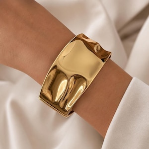 Puede incluir: Una pulsera brazalete ancha de tono dorado pulido con una superficie lisa y reflectante que presenta curvas y hendiduras orgánicas. El metal tiene un acabado de alto brillo que capta la luz, creando reflejos variados sobre su forma irregular.