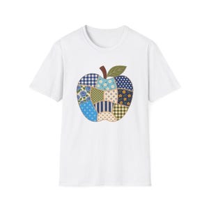 Op de afbeelding: Een wit t-shirt met korte mouwen en een centrale appelafbeelding die is samengesteld uit verschillende patchwork-stofpatronen. Het appelontwerp bevat blauwe, groene en crèmekleurige tinten met bloemen-, ruit-, stippen- en streepmotieven.