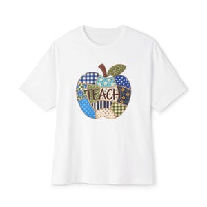 May include: A white short-sleeved t-shirt featuring a central graphic of an apple composed of various blue, green, and cream-coloured fabric patches with floral, gingham, and polka dot patterns. The word TEACH is printed in brown across the apple.