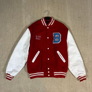 Peut inclure: Une veste universitaire rouge à manches blanches, arborant une grande lettre B bleue sur la poitrine gauche et le texte brodé U.S.I. Bruins sur la droite. La veste présente une fermeture à boutons-pression, des poignets et un col côtelés rayés.