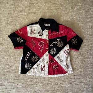 Könnte beinhalten: Ein kurzärmeliges Hemd mit Knopfleiste und einem Patchwork-Design in Rot, Schwarz und Weiß. Das Kleidungsstück ist mit verschiedenen gestickten Mustern verziert, darunter Blumenmotive und geometrische Formen, und hat einen schwarzen Kragen und goldene Knöpfe.
