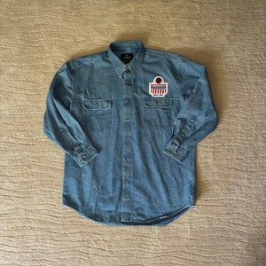 Puede incluir: Una camisa de manga larga en mezclilla azul con cuello puntiagudo, dos bolsillos en el pecho con solapas abotonadas y un parche rectangular en el pecho izquierdo. El parche muestra el texto Smith & Wesson y Since 1852 con un diseño de bandera.