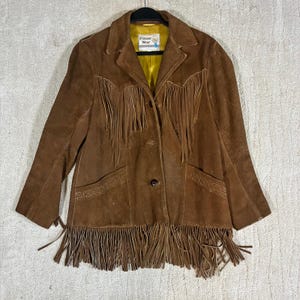 May include: A brown suede jacket featuring long fringe detailing across the chest and hemline. The jacket has a notched collar, three front buttons, and two pockets with decorative stitching. A bright yellow lining is visible, and a Pioneer Wear label.