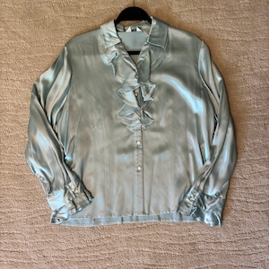 Op de afbeelding: Een blouse met lange mouwen en knoopsluiting in een bleke saliegroene kleur met een glanzende, satijnachtige afwerking. Het kledingstuk heeft een opvallende ruchestrook langs het middenvoor, een klassieke kraag en manchetten met knopen en ruches.