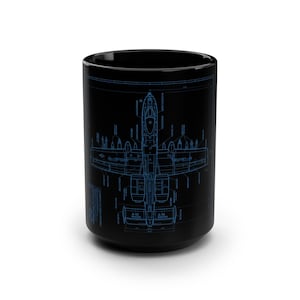 May include: A black ceramic mug featuring a detailed blue technical blueprint illustration of an A-10 Thunderbolt aircraft. The design includes precise line drawings of the plane from a top-down perspective, complete with technical labels and text.