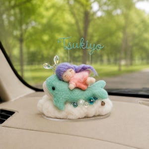 May include: A needle-felted figurine featuring a small person with purple hair sleeping on a mint green whale, resting on a white cloud base. A wire topper displays the word Tsukiyo in cursive script, and clear crystal accents are attached.