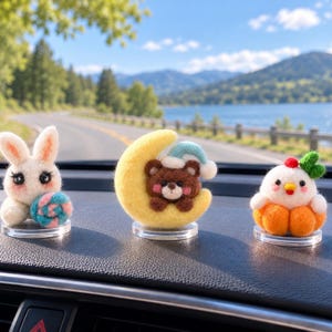 Peut inclure: Trois figurines en laine feutrée sur des bases circulaires transparentes : un lapin blanc tenant une sucette rose et bleue, un ours brun portant un bonnet bleu et blanc sur un croissant de lune jaune, et un poussin blanc avec des feuilles vertes.