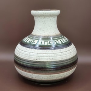 Könnte beinhalten: Eine Keramikvase mit bauchigem Körper und engem Hals, die eine gesprenkelte, cremeweiße Glasur aufweist. Die Oberfläche ist mit horizontalen Streifen in Dunkelgrün, Braun und Creme verziert, sowie einem Band mit weißer Schrift an der Schulter.