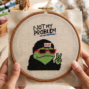 May include: A circular wooden embroidery hoop containing a piece of beige linen fabric with a cross-stitch design. The embroidery features a green frog wearing a black hoodie, red sunglasses, and a hat with the text me, making a peace sign.