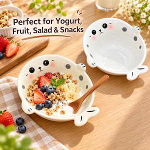 Könnte beinhalten: Zwei weiße Keramikschalen in Robbenform mit schwarzen Augen, Nasen und grauen Punkten. Eine Schale ist leer, die andere enthält Joghurt, Granola, Bananenscheiben, Erdbeeren, Blaubeeren und einen Minzzweig mit einem Holzlöffel.
