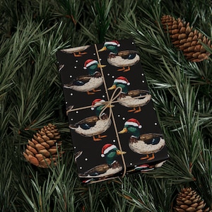 Puede incluir: Una caja de regalo rectangular envuelta en papel negro con un patrón repetitivo de patos azulones que llevan gorros de Papá Noel rojos y blancos, acentuado con pequeños puntos blancos y atado con un cordel fino de color marrón claro.